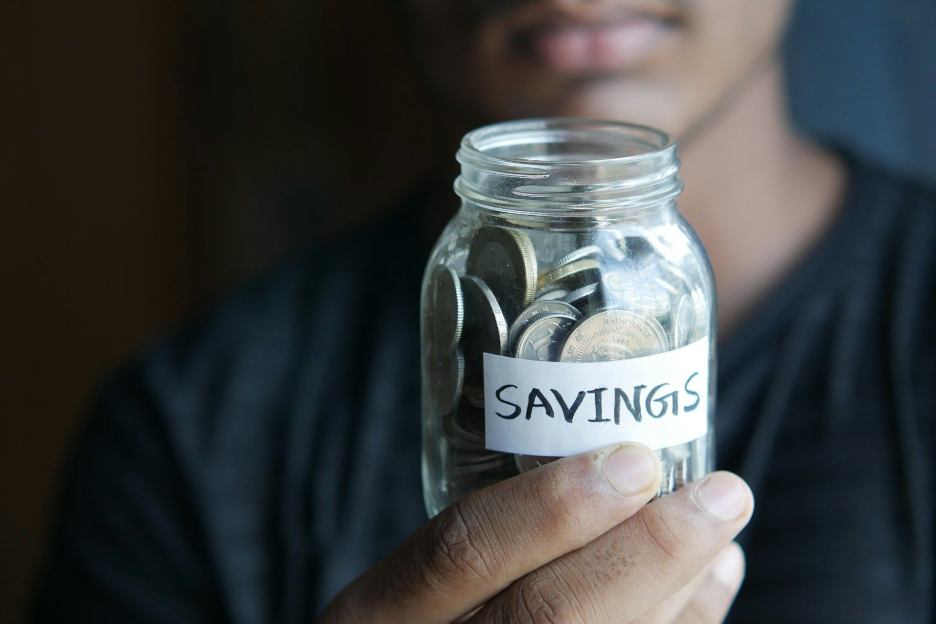 A close-up shot of a person's hands placing a coin into a jar labeled "Retirement." The image has a clean, focused feel, symbolising the act of consistent saving for a long-term goal.